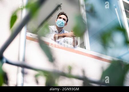 Young businessman wearing protective face mask standing with hands clasped at window Stock Photo