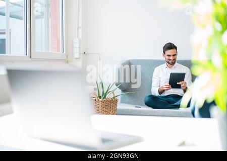 Smiling young businessman using digital tablet while sitting on sofa in office Stock Photo