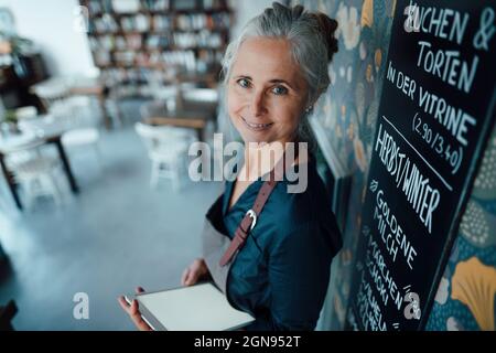 Smiling female cafe owner wearing apron leaning on blackboard in coffee shop Stock Photo