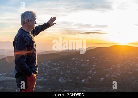 Senior man shielding eyes while standing on mountain during sunset Stock Photo