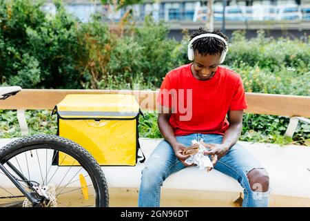 Happy delivery man listening music through wireless headphones while eating food on bench Stock Photo