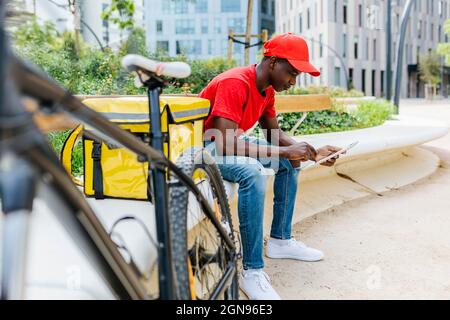 Young delivery man using digital tablet while sitting by package on bench Stock Photo