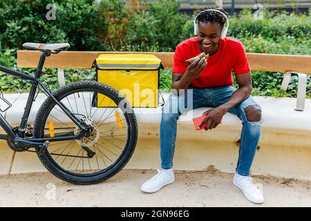 Smiling young delivery man eating food while sitting by bicycle on bench Stock Photo