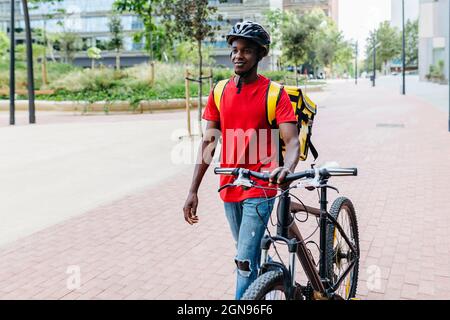 Smiling delivery man with bicycle walking on footpath Stock Photo