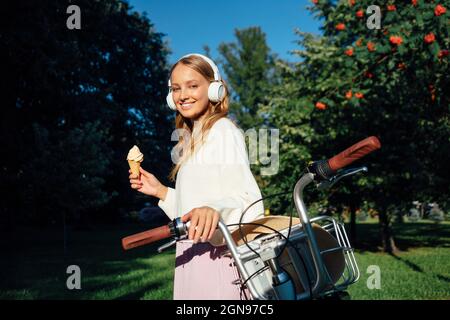 Young woman wearing headphones holding ice cream by bicycle at park during sunny day Stock Photo