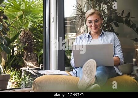 Smiling female freelancer using laptop while sitting by window Stock Photo
