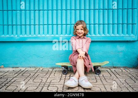 Smiling girl sitting with legs crossed at knee on skateboard in front of blue wall Stock Photo