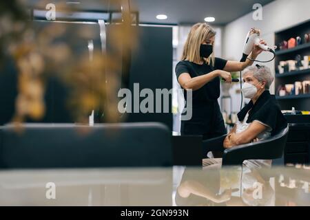 Female hairdresser with face mask blow drying customer's hair in salon Stock Photo