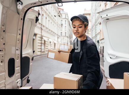 Woman courier in uniform carefully unloading cardboard boxes from car for delivery Stock Photo