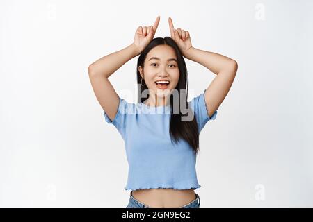 Happy asian woman pointing fingers up, showing finger horns gesture on head, smiling and looking enthusiastic, white background Stock Photo