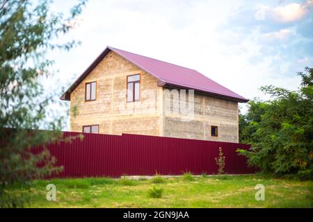 A house with a crimson roof behind a fence in a village in summer. Construction of suburban housing. Stock Photo