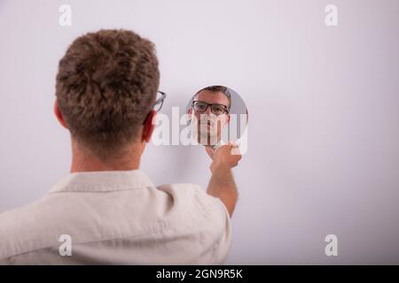 Serious caucasian man looks at himself in the mirror and reflects on life. Pensive person with eyeglasses. Guy isolated on white background Stock Photo