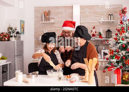 Happy family cooking homemade gingerbread dessert making traditional dough celebrating christmas holiday together. Grandchild enjoying winter season cooking delicious cookies in xmas decorated kitchen Stock Photo