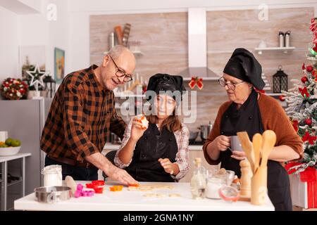 Happy family cooking traditional gingerbread dessert making dough using baking tree shape in xmas decorated kitchen. Grandchild celebrating christmas holiday together preparing homemade cookies Stock Photo