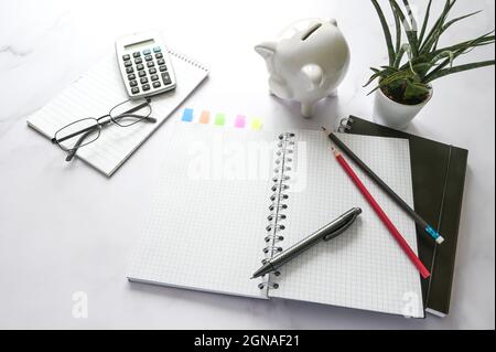 Office desk with a blank notebook, piggy bank, calculator and stationary supplies on a marble surface, copy space, selected focus, narrow depth of fie Stock Photo