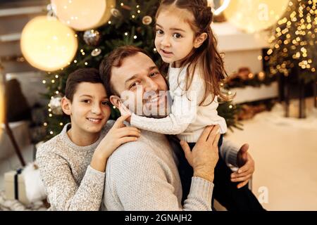 Portrait of caring father with little kids sitting near Christmas tree, smiling. Happy man with adorable daughter and joyful son celebrate New Year at Stock Photo