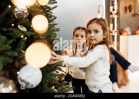Portrait of beautiful sisters decorating Christmas tree, smiling, spending time together. Adorable little girls celebrating winter holidays, having Stock Photo