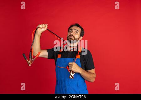 Portrait of young bearded man, male auto mechanic or fitter wearing blue work dungarees isolated over red studio background. Concept of facial Stock Photo
