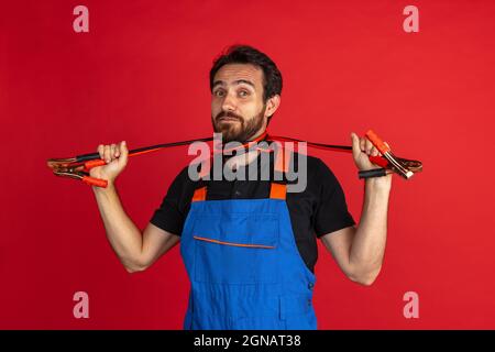 Portrait of young bearded man, male auto mechanic or fitter wearing blue work dungarees isolated over red studio background. Concept of facial Stock Photo