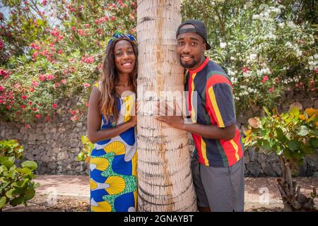 Happy laughing playful african millennial spouses posing hugging a palm tree, smiling and looking at camera, siblings, brother and sister posing Stock Photo