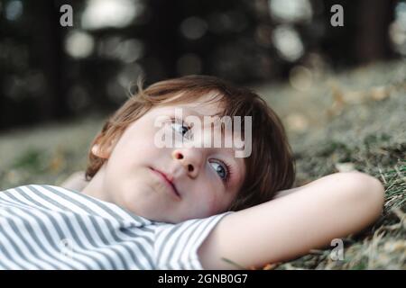 Kid lying on his back in the grass in park. Boy chilling on green lawn. Child relaxing outside, resting his head on garden meadow Stock Photo