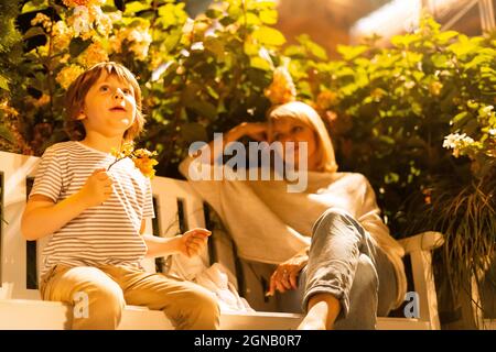 Boy and his mom sitting on the bench in city park. Son and mother having fun on log seat with bushes in the background. Autumn park family leisure Stock Photo