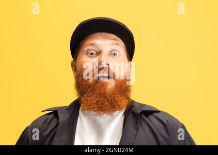Close-up portrait of stylish red-bearded man, barber in black cap looking at camera isolated over yellow background. Concept of emotions, feelings. Stock Photo