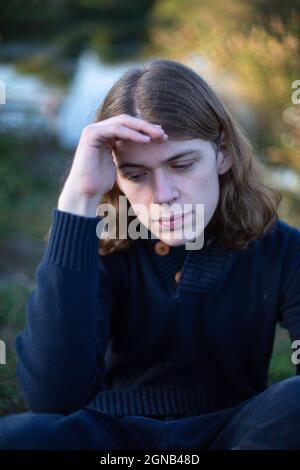Sad young european man with long hair with lake behind Stock Photo