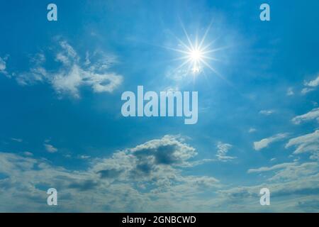 The midday sun shines brightly high in the blue sky. Azure sky with small white clouds on a sunny day. Sun in the sky for weather forecast background. Stock Photo