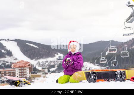 Cute adorable preschooler caucasian kid girl portrait little girl in santa hat and snowboard enjoy winter sport activities. Stock Photo