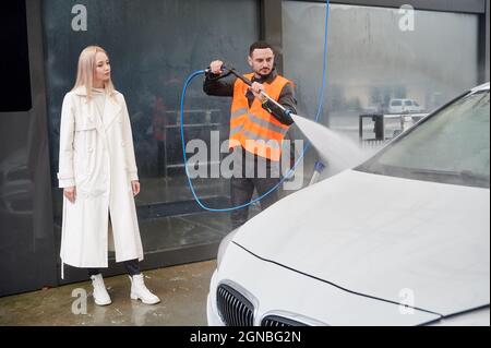 Young man washing car on carwash station, wearing orange vest. Handsome worker cleaning automobile, using high pressure water. Beautiful blonde woman client looking on the white car. Stock Photo