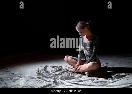 Pretty young female in black bodysuit sitting on floor with crossed legs having rest. female adult ballet dancer performer in flour. lady dancer with Stock Photo