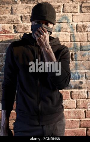 african american man in mask standing by brick wall, hooligan in black wear. black guy pose a danger for society, waiting for victim in dark alley, co Stock Photo