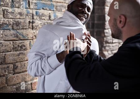 Street theif commits robbery attack on african man, at day time. Young frightened black man is being assaulted by dangerous angry man in alley. Crime Stock Photo