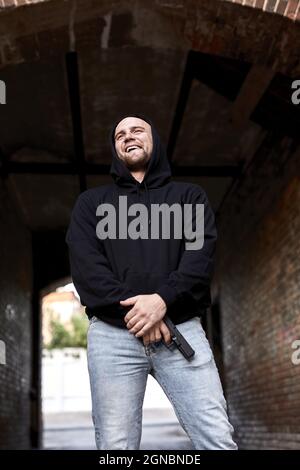 Young Cruel Hooligan man in the hood with gun standing at street alley, smiling caucasian male is waiting for victim, commit a crime. Outdoor portrait Stock Photo
