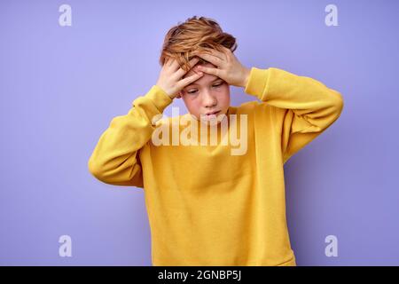 Teenage boy is suffering from headache, touching head, isolated on purple background. Portrait of young boy in casual yellow shirt desperate and stres Stock Photo