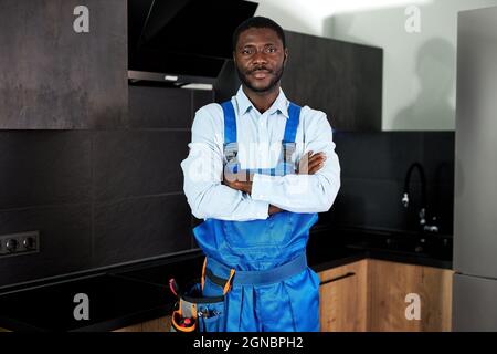 Serious Handsome Young African Handyman Plumber In Blue Uniform. Young Skilled Handyman In Overall Standing At Home In Kitchen Posing Looking At Camer Stock Photo