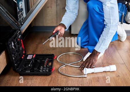 Unrecognizable Professional Handyman Of African American Appearance In Blue Overalls Repairing Dishwasher, Using Instruments Opening Toolbox, In Kitch Stock Photo