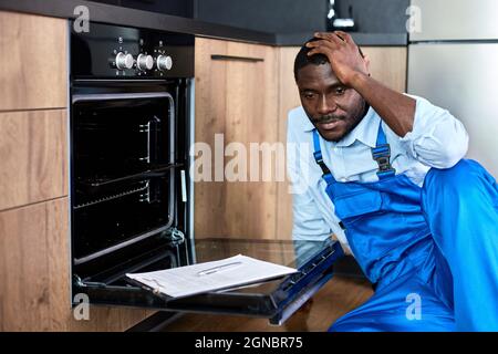 Upset repairman in uniform having troubles in work while repairing oven, sit on floor touching head, thinking about how to solve problem, how to repai Stock Photo