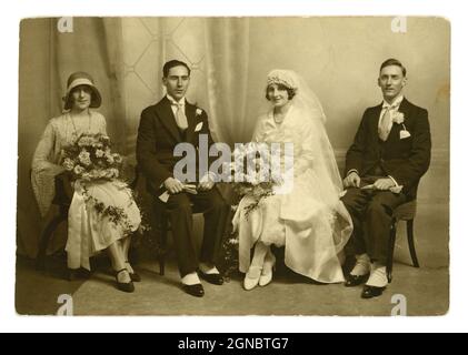 Stunning original classic 1920's, flapper era, wedding group portrait photo, beautiful bride wearing a long veil, stunning headpiece. sitting next to the groom and best man wearing matching suits with winged shirt collars, spats. The bridesmaid or maid of honour is wearing a fashionable cloche hat, circa 1924, U.K. Stock Photo