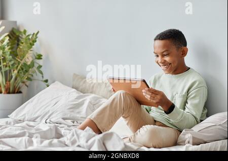 Full length portrait of smiling African-American teenager using tablet while sitting on bed in cozy room, copy space Stock Photo