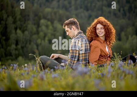 Lovely Caucasian Couple Sit On Grass Relaxing In Countryside, In Contemplation, Enjoying Nature, In Casual Wear, Redhead Lady With Curly Hair And Man Stock Photo