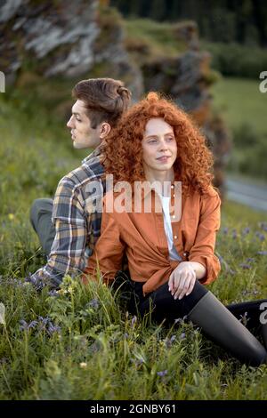 Dreamy Caucasian Couple Sit On Grass Relaxing In Countryside, In Contemplation, Enjoying Nature, In Casual Wear, Redhead Lady With Curly Hair And Man Stock Photo