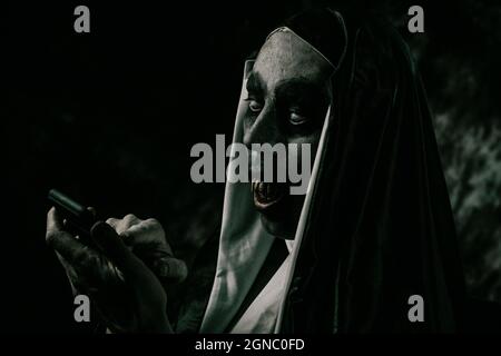 closeup of a scary evil nun, with bloody teeth, in a typical black and white habit, using her smartphone Stock Photo