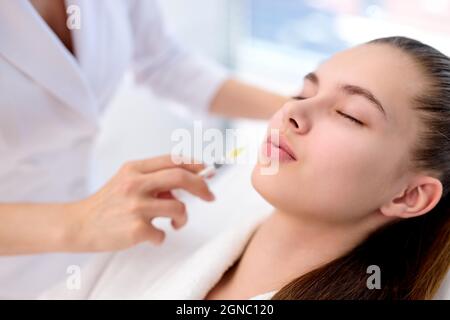 female cosmetologist making botox injection in female lips, holding syringe. young beautiful caucasian woman relaxing on couch in bathrobe. Aesthetic Stock Photo