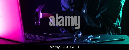 Criminal hacker in a hood in black clothes and balaclava destroys a laptop with a hammer on a black background Stock Photo