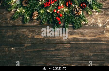 Christmas and new year card with fir branches, winter berries, pine cones and garland on brown wooden background Stock Photo