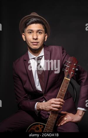 Caucasian man from Spain in suit and hat holding a guitar, sitting and looking at camera Stock Photo