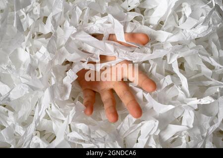 Hand reaches out from heap of shredded paper, man sinking in pile of white confetti. Somebody drowning in paper trash, garbage or box filler. Concept Stock Photo