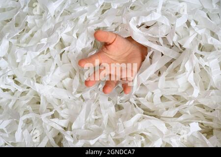 Hand reaches out from heap of shredded paper, man sinking in pile of white confetti. Somebody drowning in paper trash, garbage or box filler. Concept Stock Photo